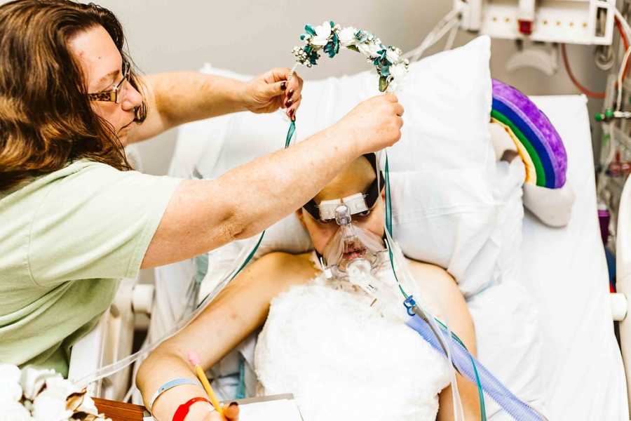 Mother places flower crown on teen daughter who lays in hospital bed wearing wedding dress