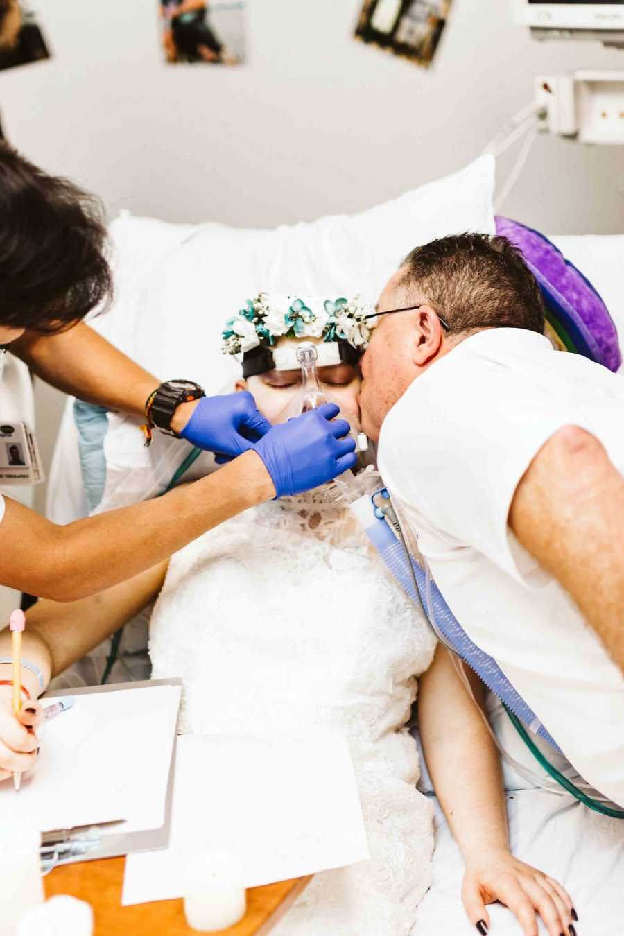 Father leans over to kiss teen daughter on cheek who is in hospital bed while nurse removes breathing tubes