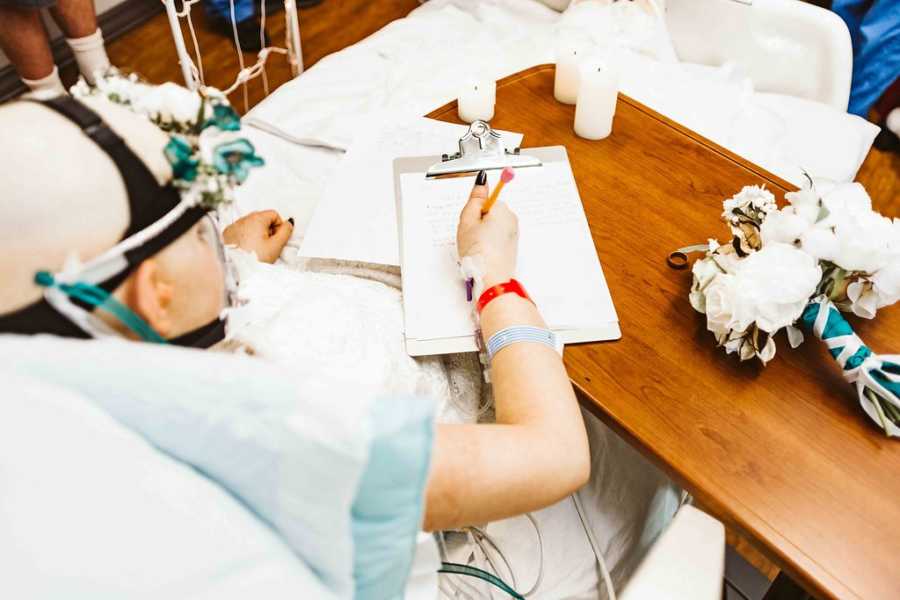 Teen with cancer sits up in hospital bed writing on clip bored with flower bouquet beside it