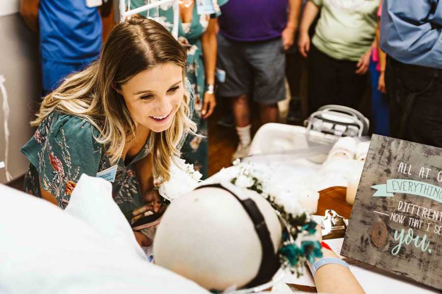 Young woman smiles while leaning over hospital bed where teen with cancer lays before her wedding