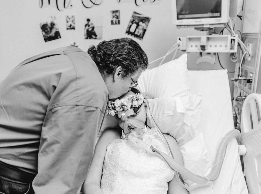 Father leans over to kiss head of teen daughter with cancer in hospital bed wearing wedding dress