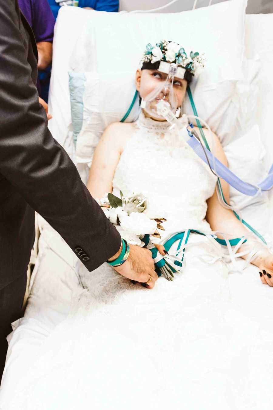 Teen bride with cancer sits in hospital in wedding gown looking up at groom