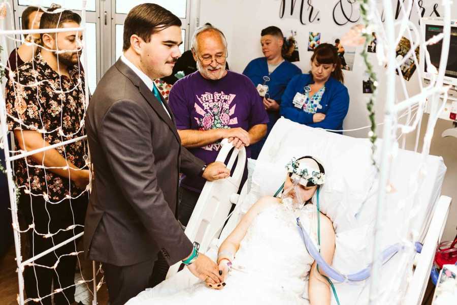 Teen bride with cancer lies in hospital bed holding hands with groom while people watch 