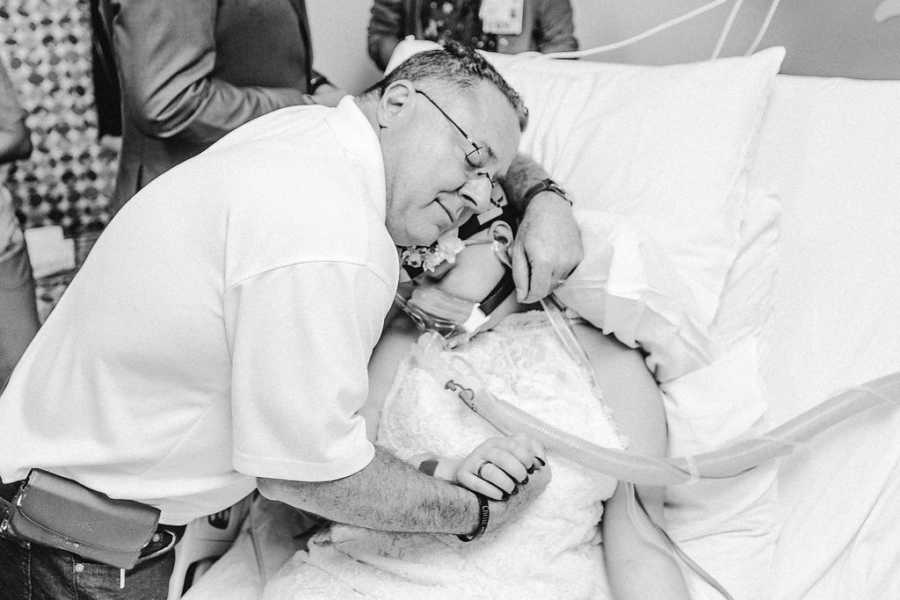 Man leans over hospital bed hugging teen bride with cancer