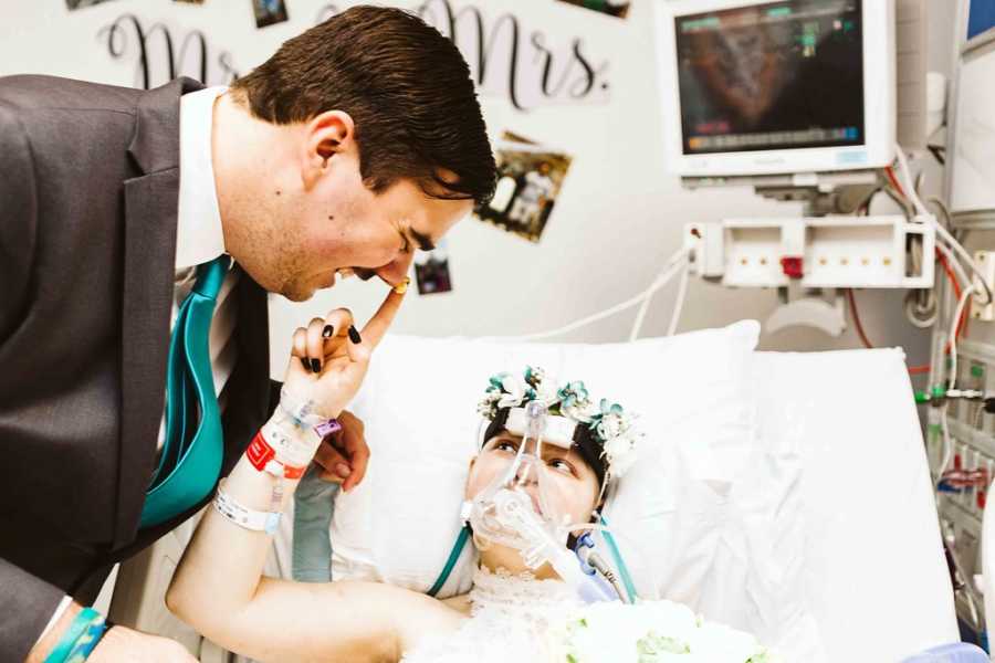 Teen bride with cancer lays in hospital bed putting frosting from wedding cake on groom's nose