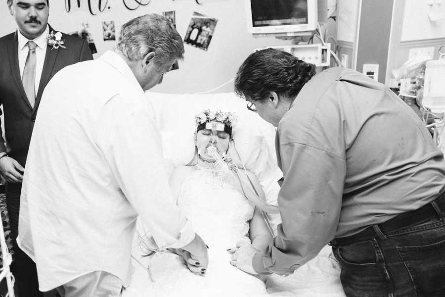 Teen bride with cancer lays in hospital bed holding hands of two men while groom stands beside her