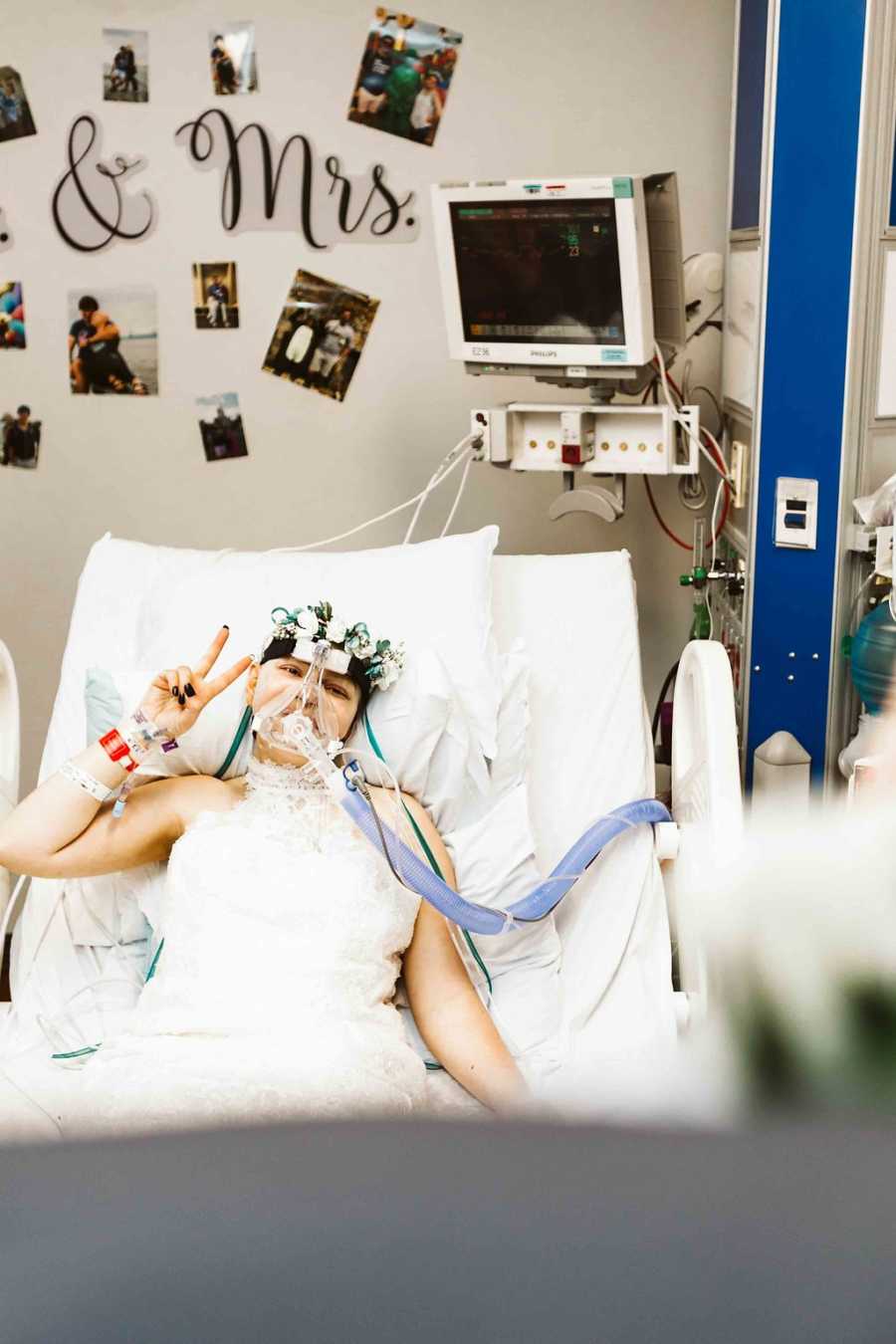 Teen bride with cancer lying in hospital bed holding up peace sign with pictures on wall behind her