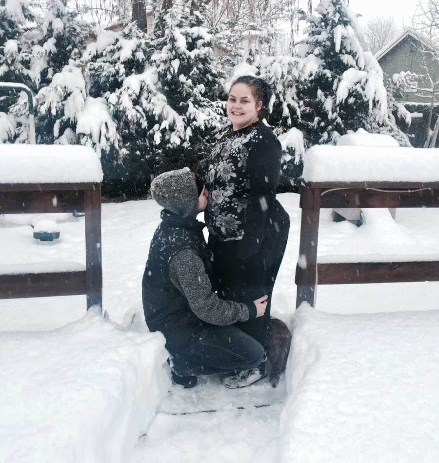 Pregnant woman stands in snow while husband crouches down kissing her stomach