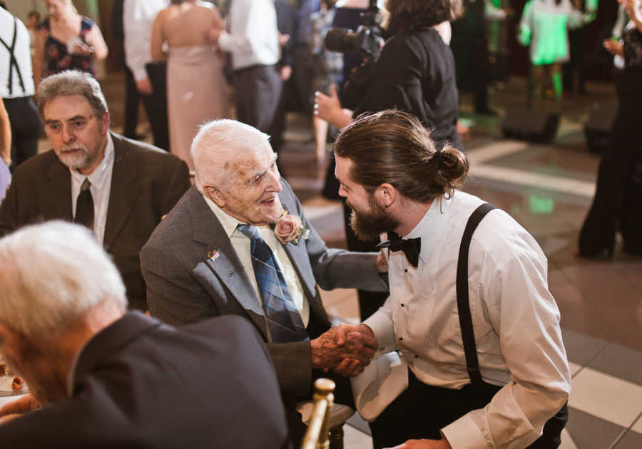 Groom squats down to shake bride's grandfather's hand as he sits at table