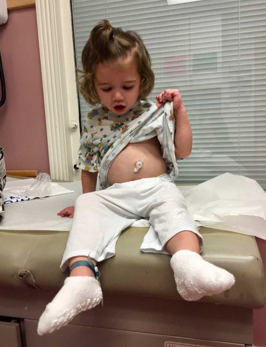 Young girl who had shaken baby syndrome holds up shirt and looks at G tube in her stomach