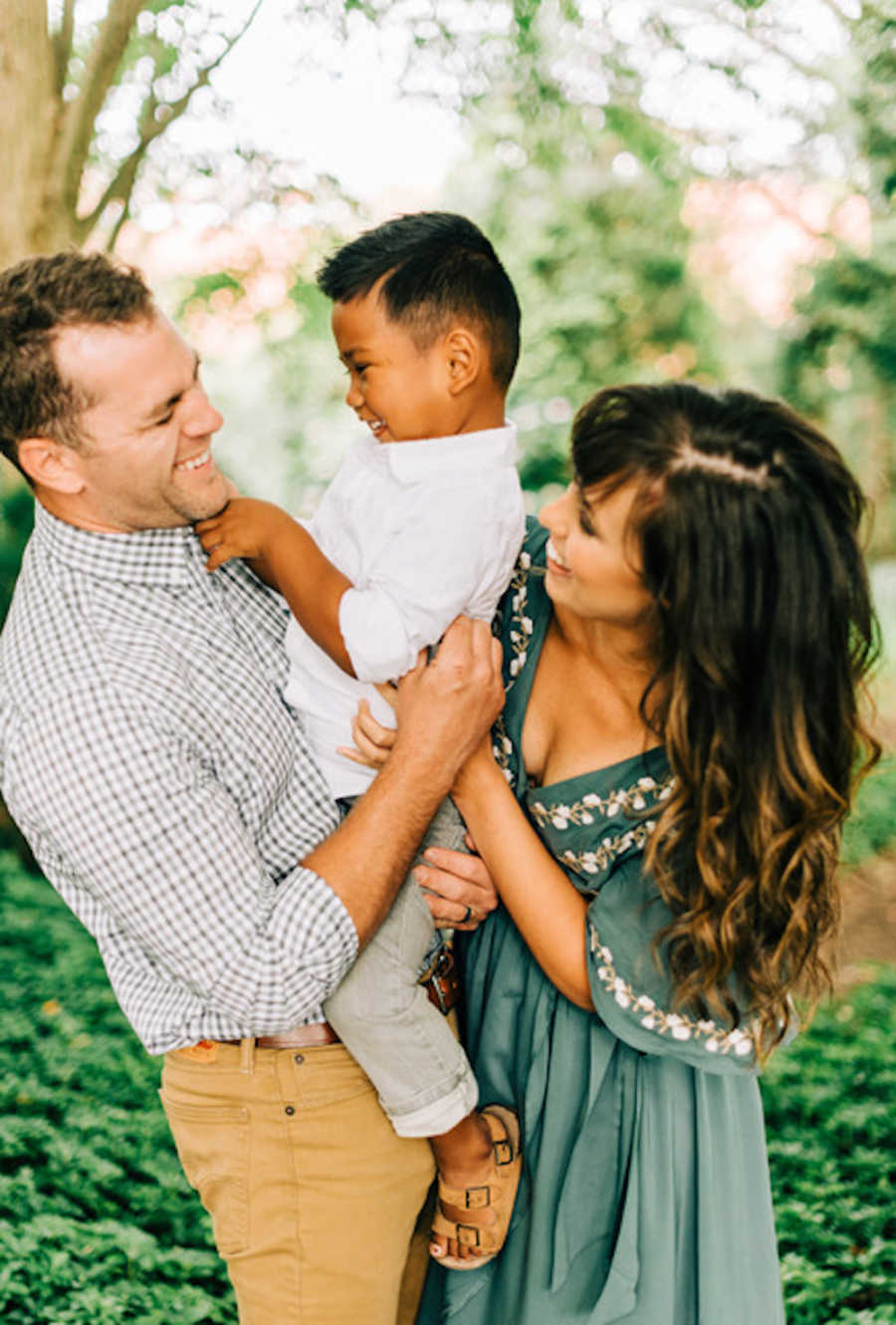 Father smiles as he holds adopted son and wife stands beside them smiling