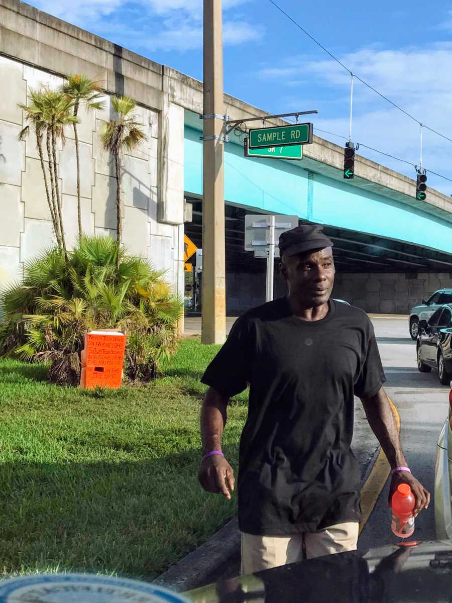 Homeless man standing beside cars on road holding a red gatorade