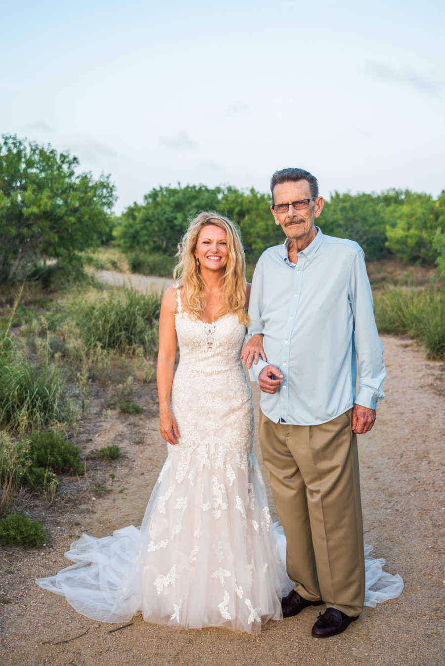Woman in wedding gown stands on dirt path arm in arm with father who has cancer