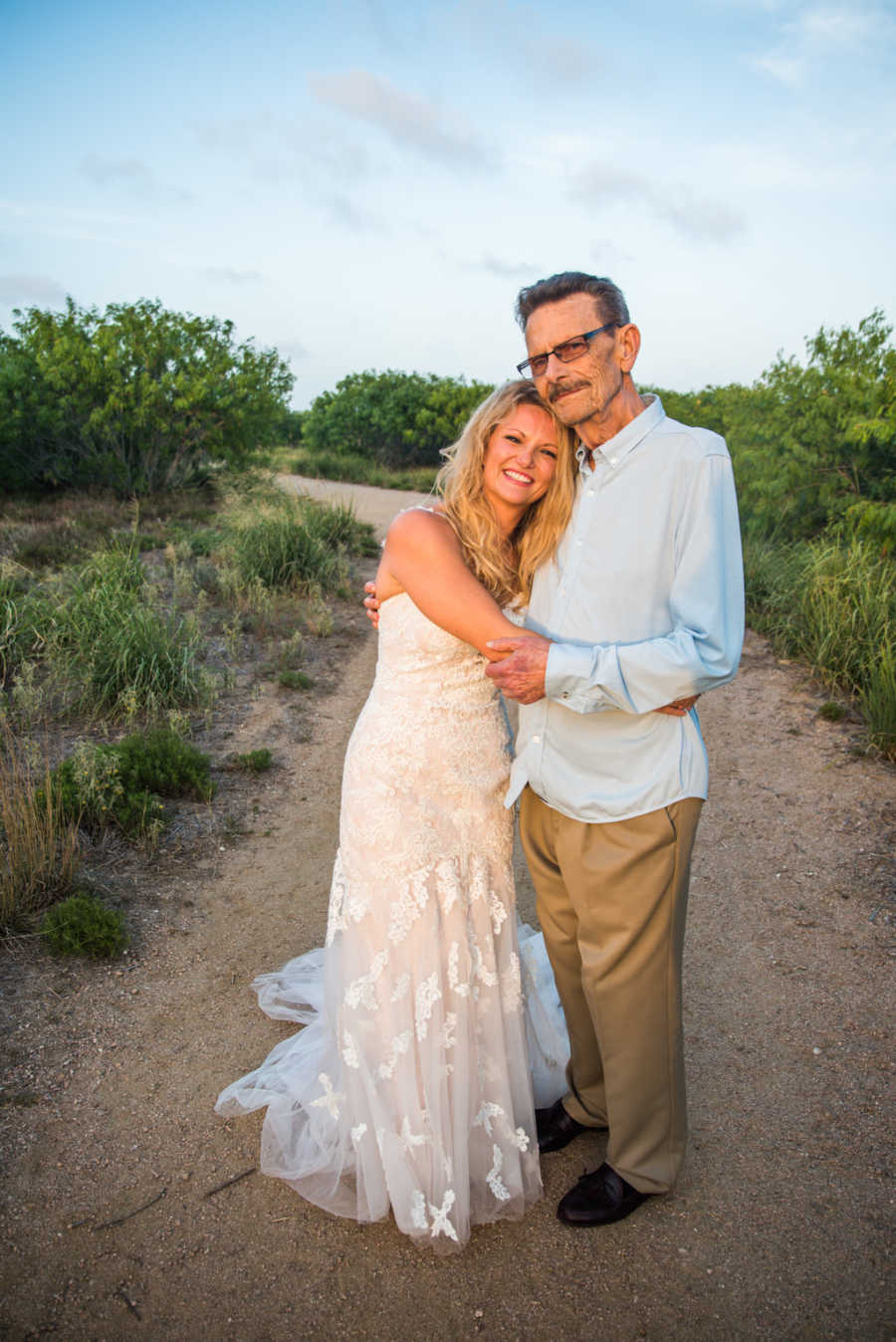 Woman in wedding gown stands leaning her head on fathers chest who has cancer 