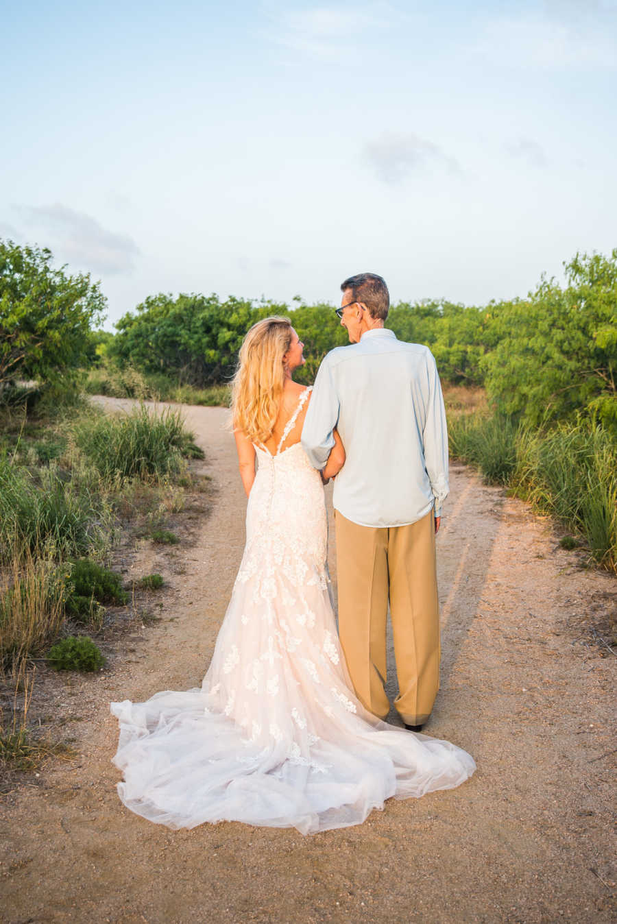 View from back of woman in wedding gown arm in arm with her father who has cancer