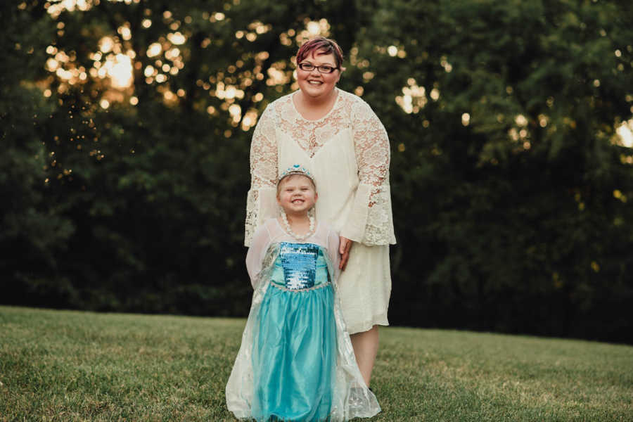 Little girl smiling in princess dress with mother in white dress standing behind her