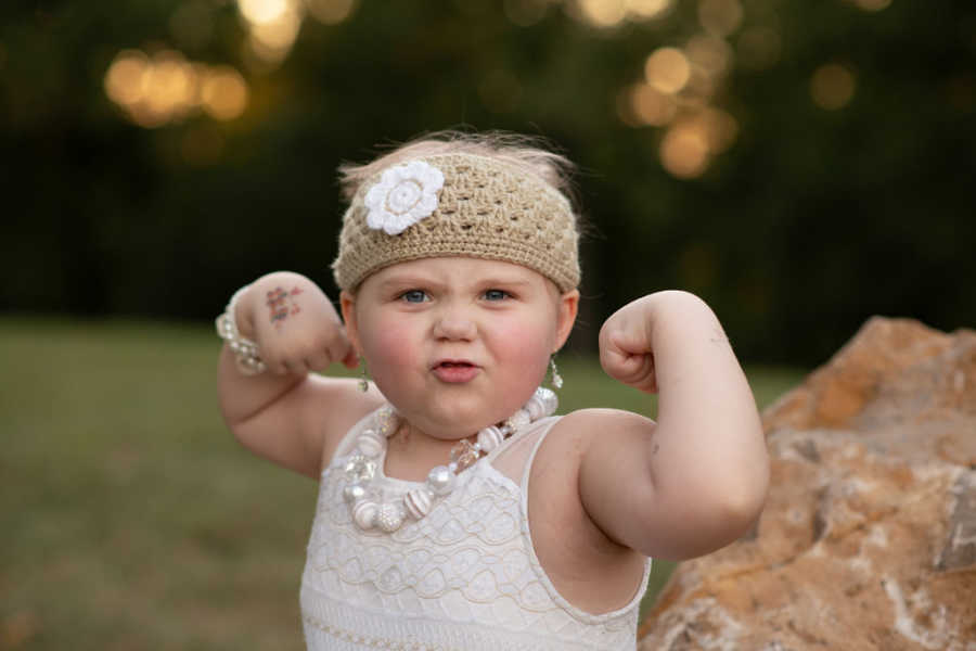 Little girl with leukemia flexing her muscles wearing a white dress and flower headband