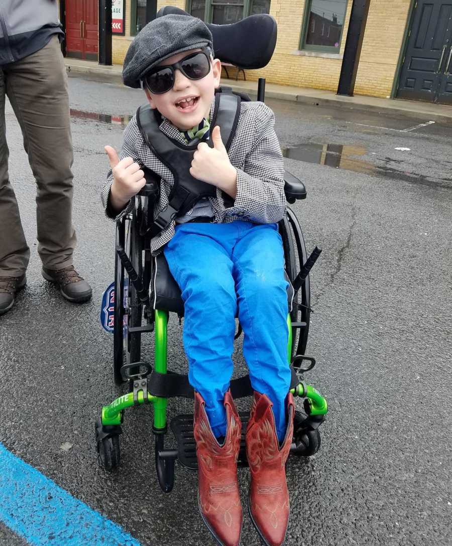 Boy with vanishing white matter disease sits in wheelchair with thumbs up wearing sunglasses and hat