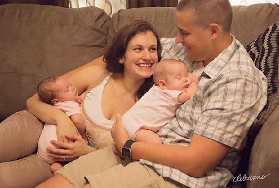 Husband and wife sitting on couch smiling at each other with twins in their lap