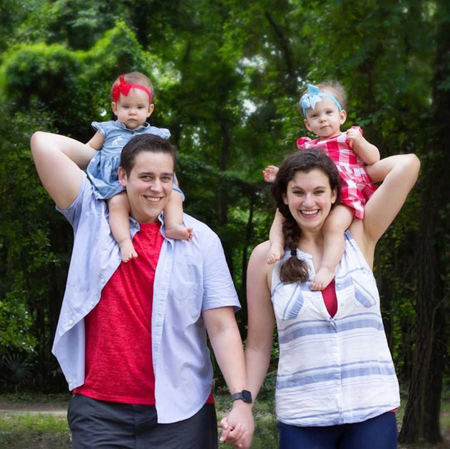 Husband and wife stand holding hands with their toddler twins on their shoulders