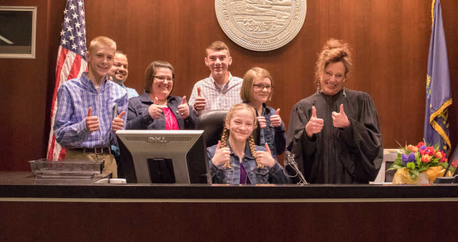 Husband and wife stand with thumbs up beside four adopted children and adoption judge