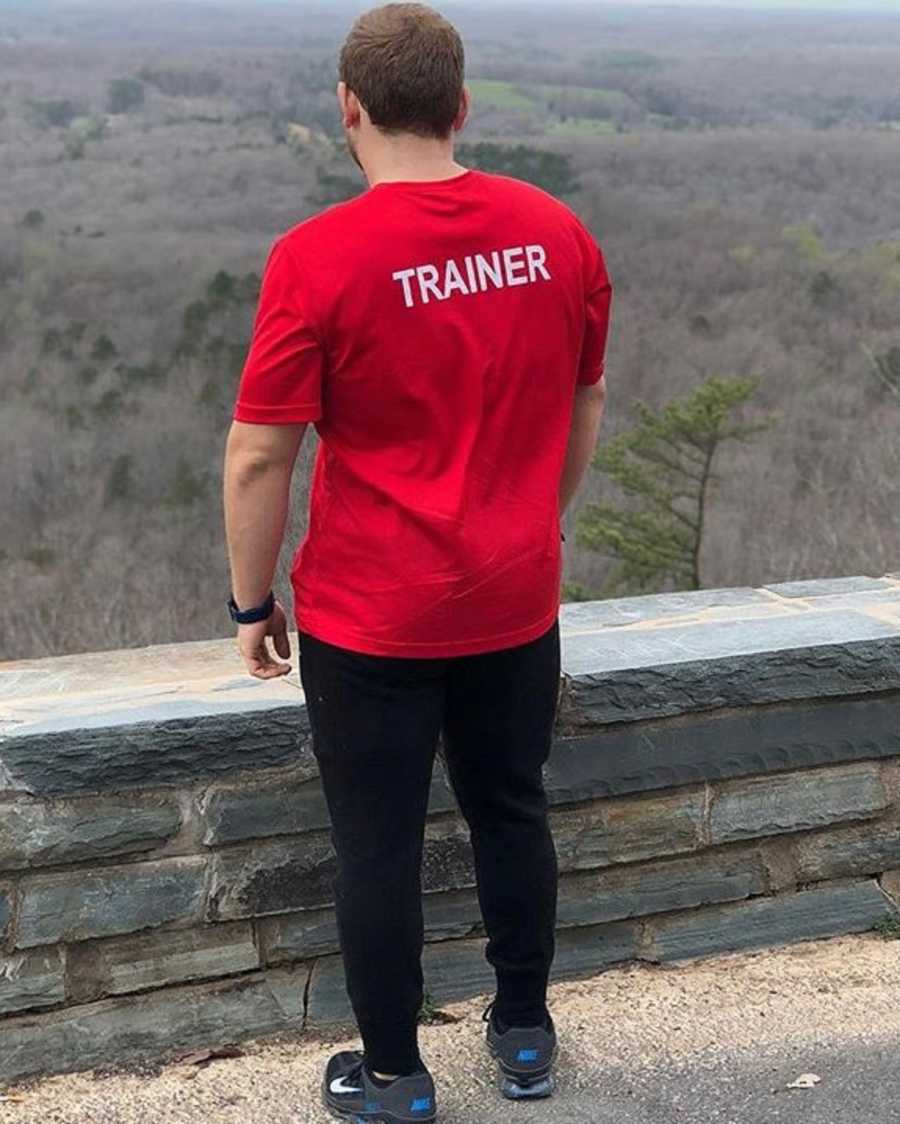 Man who once weighed 320 pounds looks out at scene in front of him wearing shirt that says, "Trainer"