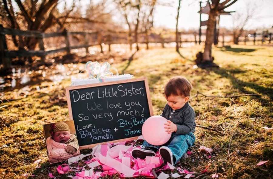 Toddler sits on ground outside with pink balloon beside chalkboard announcing birth of his sister