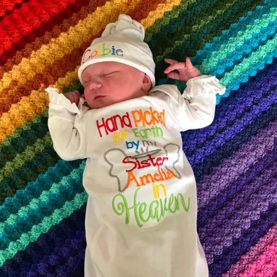 Newborn laying on rainbow blanket wearing onesie that says, "Hand picked for earth by my sister Amelia in heaven"