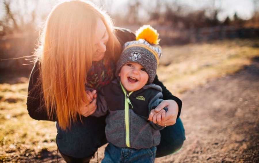 Mother who lost her first child crouches down beside toddler foster son who is smiling 