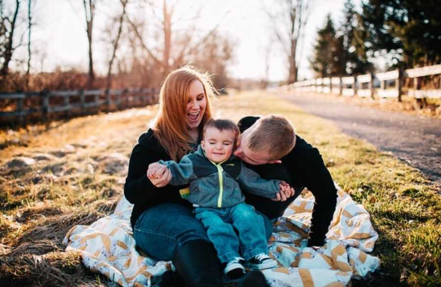 Mother sits on blanket outside with toddler foster son in her lap while husband kisses his cheek
