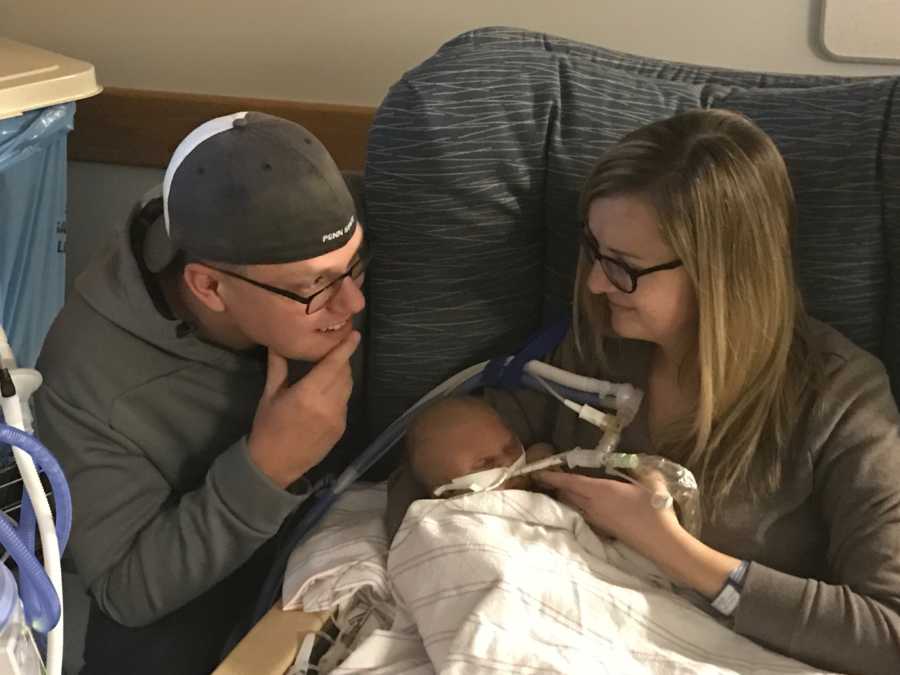Mother sits in chair holding newborn with chromosomal abnormalities while looking over at husband beside her