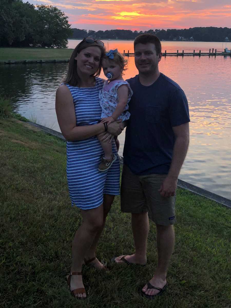 Woman holds toddler daughter beside husband who stand beside body of water