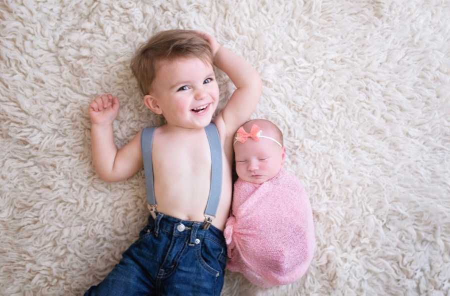 Toddler lays shirtless in suspenders and jeans on fuzzy rug with newborn sister swaddled in pink blanket beside her