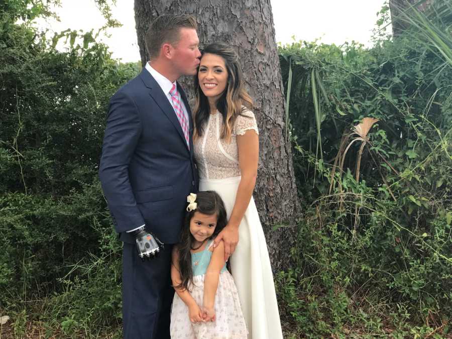 Groom kisses bride's forehead with brides daughter standing in front of them