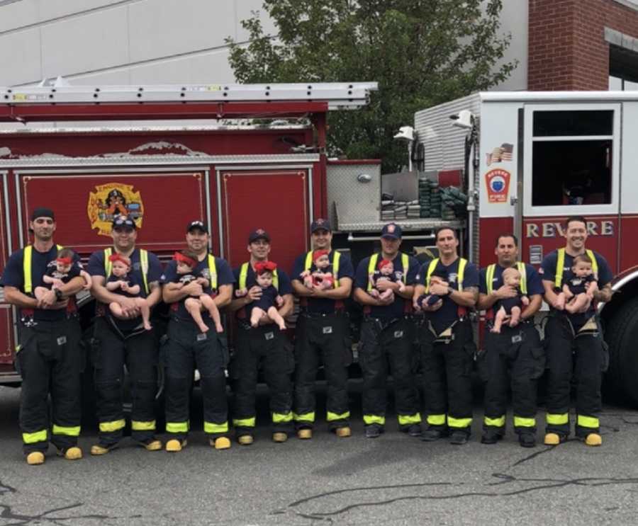 Nine firefighters stand in front of fire truck holding their newborn babies
