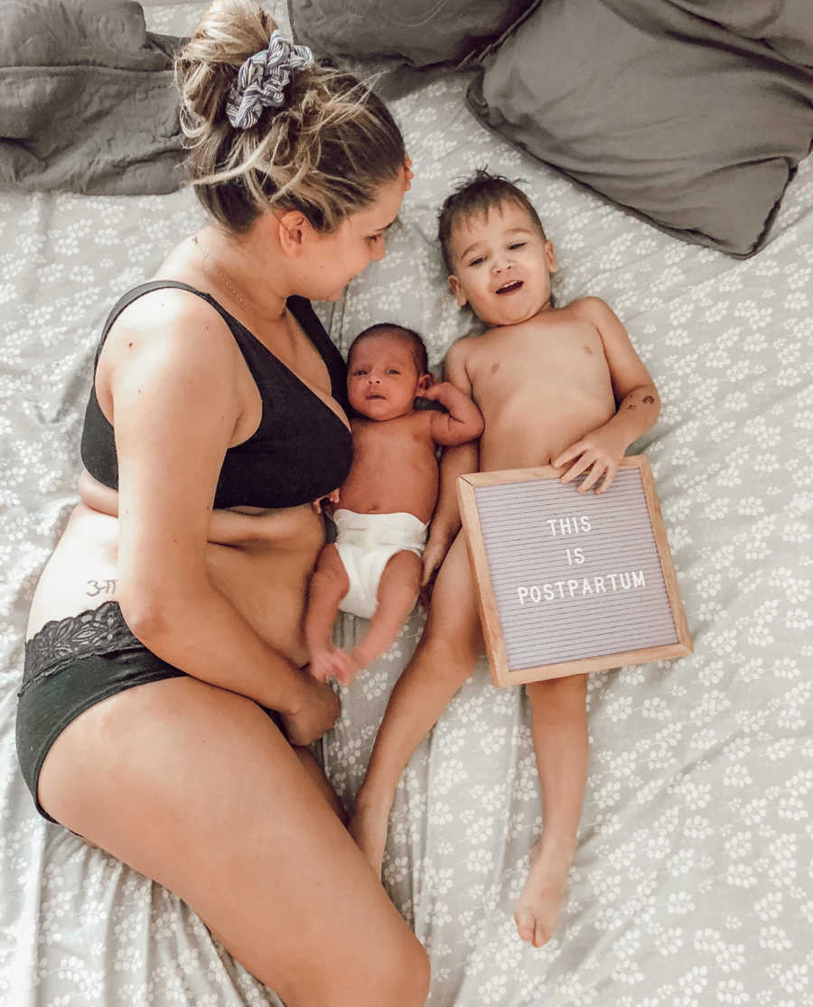 Mother lying in bed in bra and underwear beside newborn and toddler who holds sign saying, "This is postpartum"