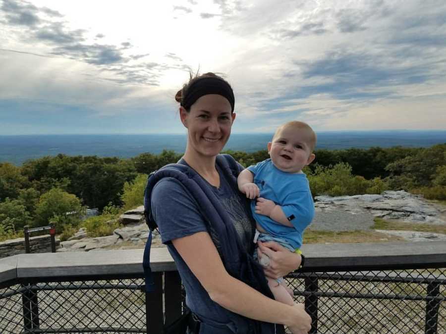 Woman smiles while holding baby son beside railing that looks out to woods and body of water