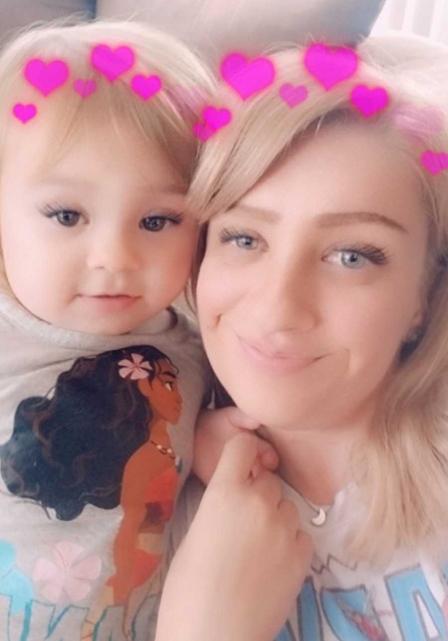 Mother smiles in selfie with toddler daughter with heart filter above their heads