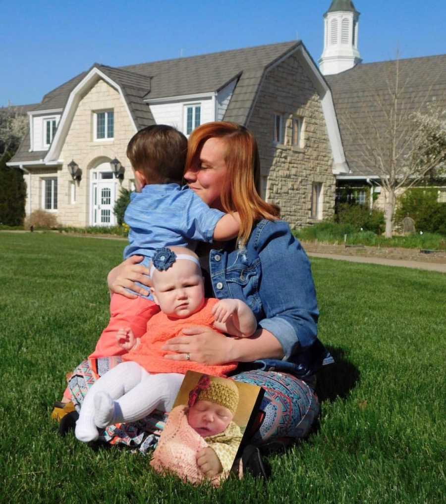 Mother sits on ground outside hugging adopted son with daughter in her lap beside picture of deceased first born