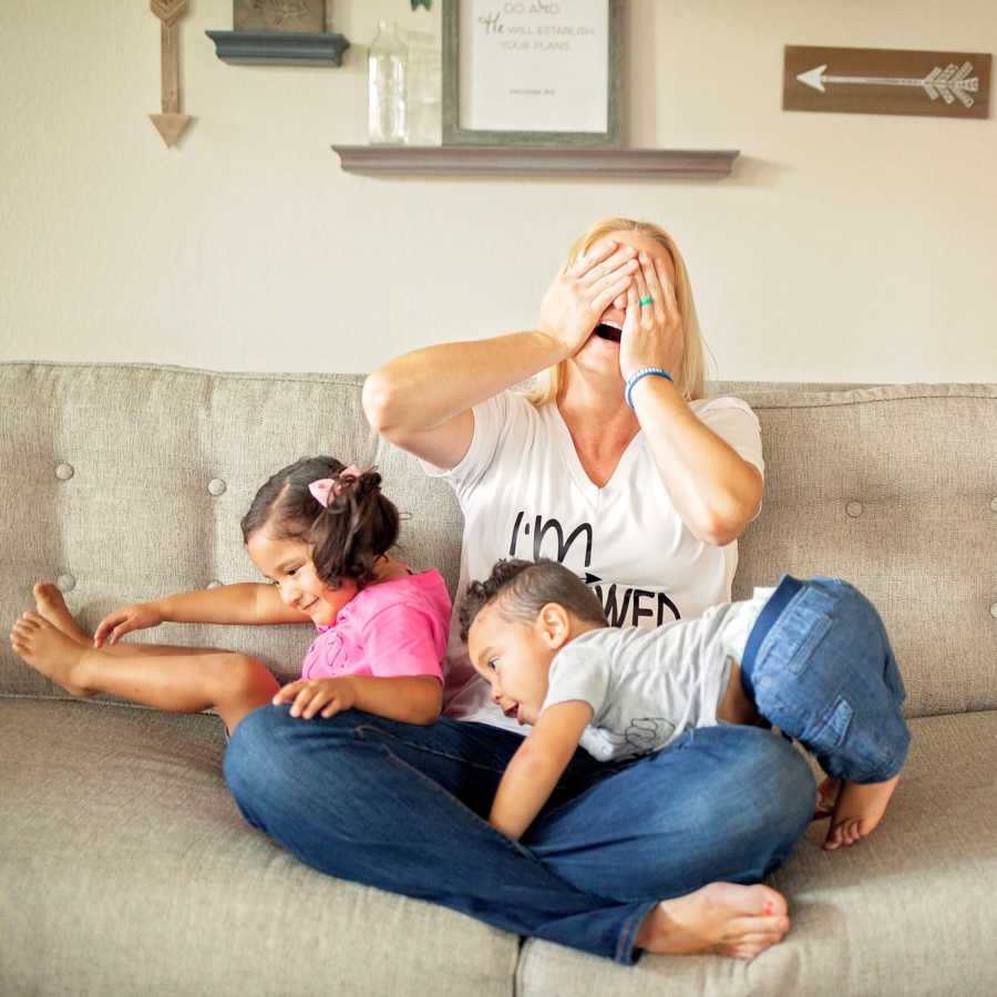 Foster mother sits on couch holding hands over her eyes with her older child and baby with no hips or femur