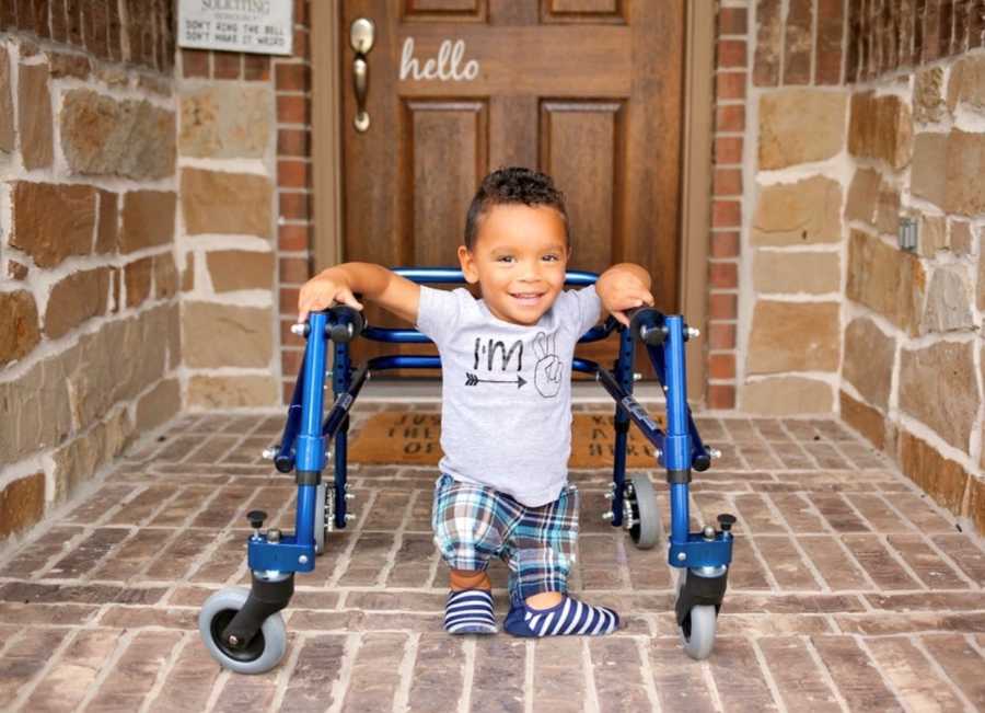 Toddler foster child born without femur or hips stands smiling on front porch with walker