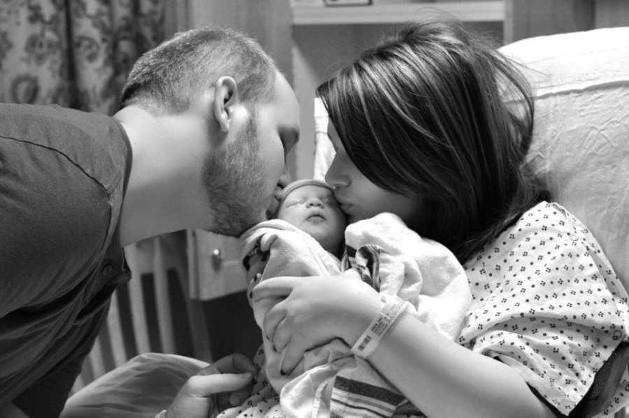 Mother holds newborn in her arms and she and her husband kiss her on either cheek