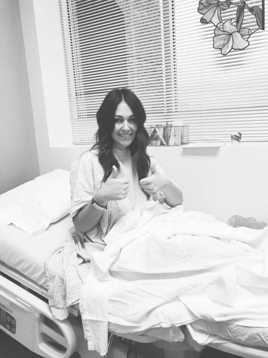 Woman who struggles with conceiving second child sits up in hospital bed with thumbs up at egg retrieval 