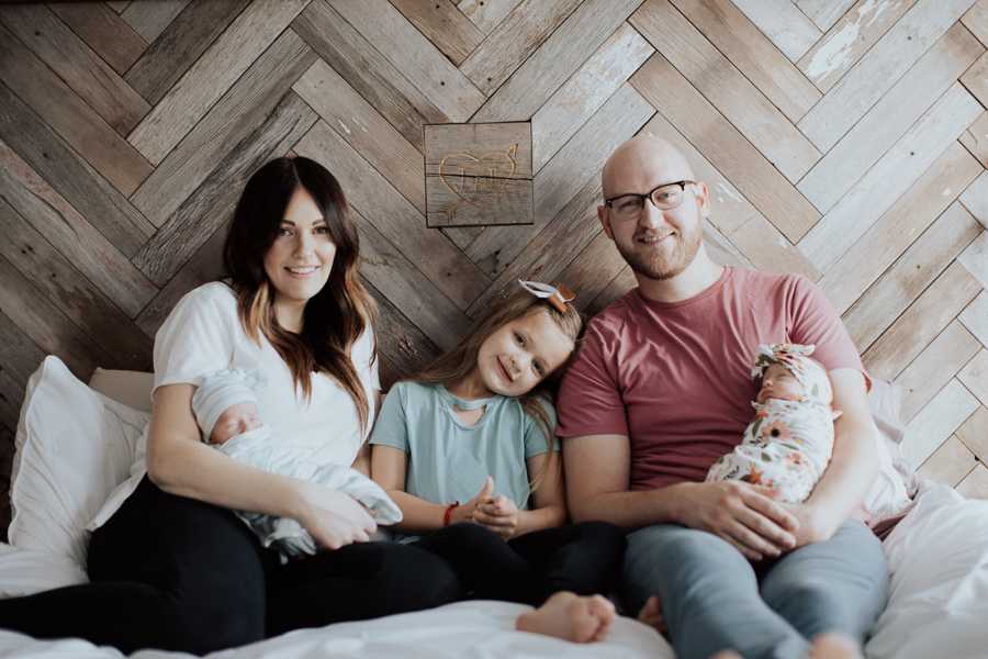 Husband and wife sit on bed holding their newborn twins with their firstborn sitting in between them