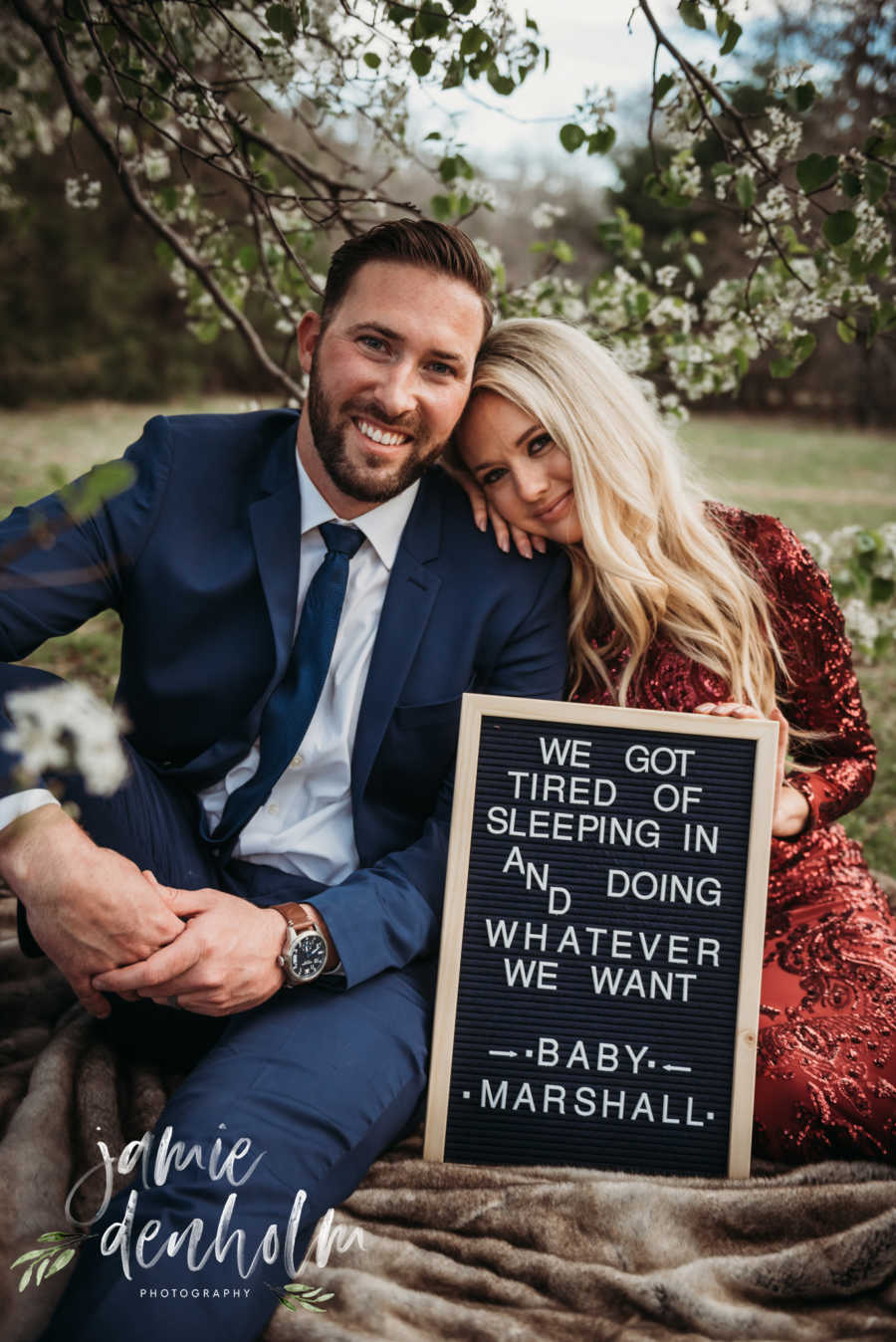Wife sits on ground beside husband resting her head on his shoulder with sign announcing pregnancy