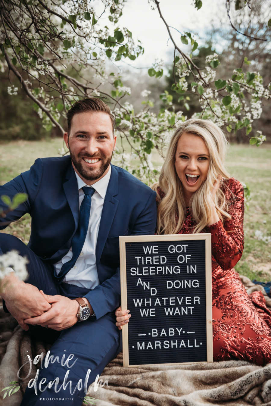 Husband and wife sit on blanket outside smiling beside sign that says, "We got tired of sleeping in..."