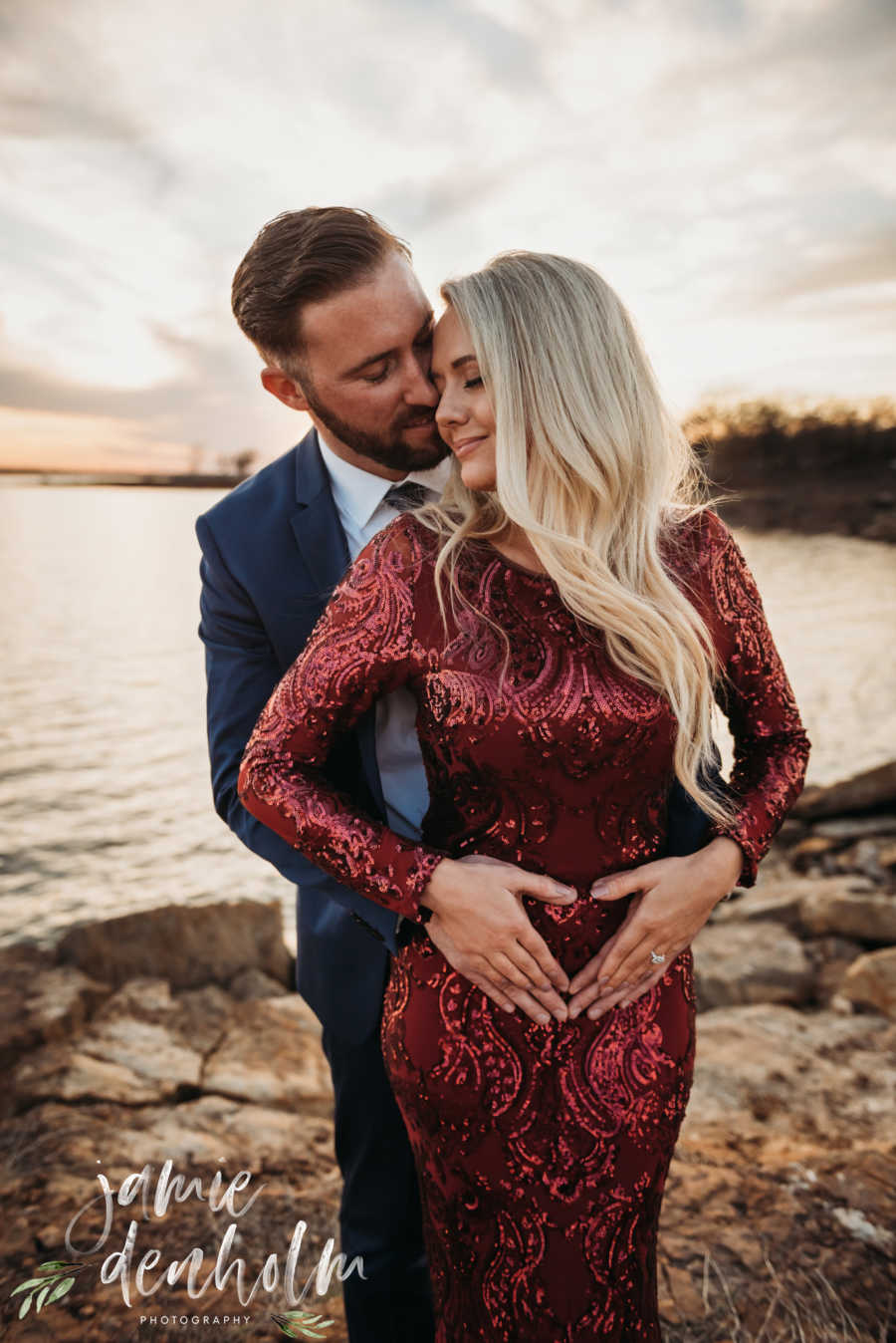 Wife stands with hands on her stomach in shape of heart with husband standing behind her with hands over hers