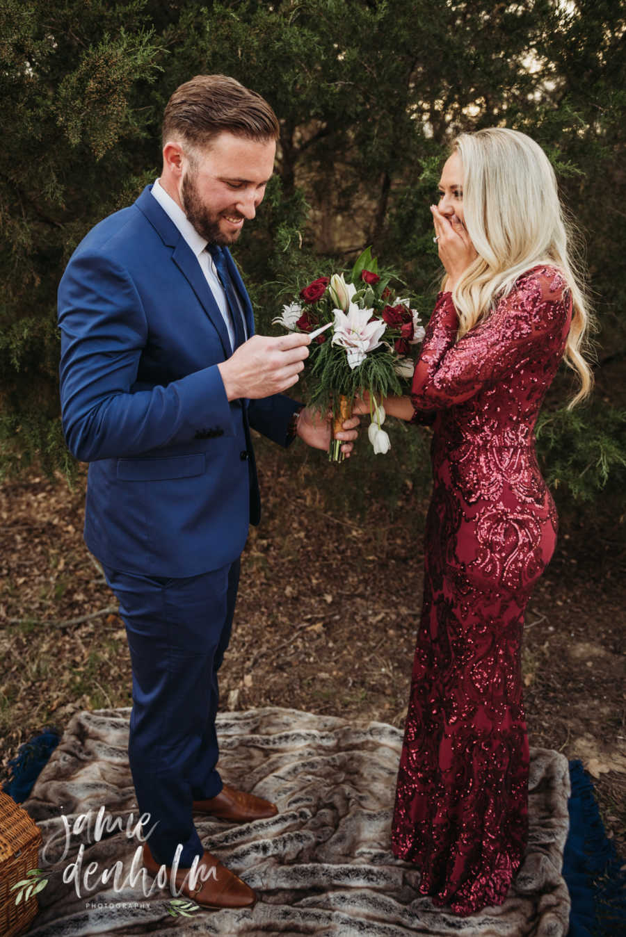 Husband smiles as he looks at pregnancy test while wife stands with hand over her mouth and bouquet of flowers