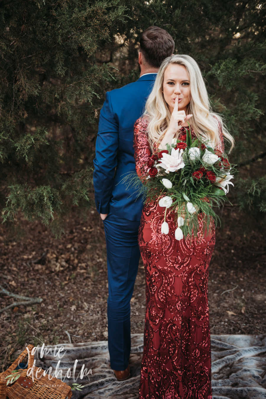 Husband and wife stand back to back while wife holds bouquet of flowers and holding finger to lip