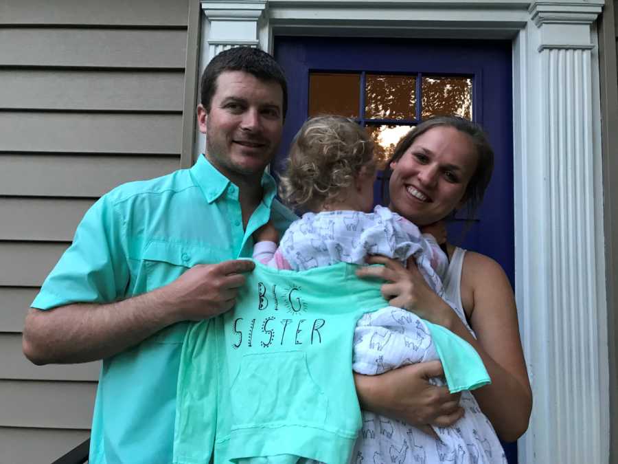 Mother holding toddler daughter beside husband while they hold sweatshirt that says, "Big sister"